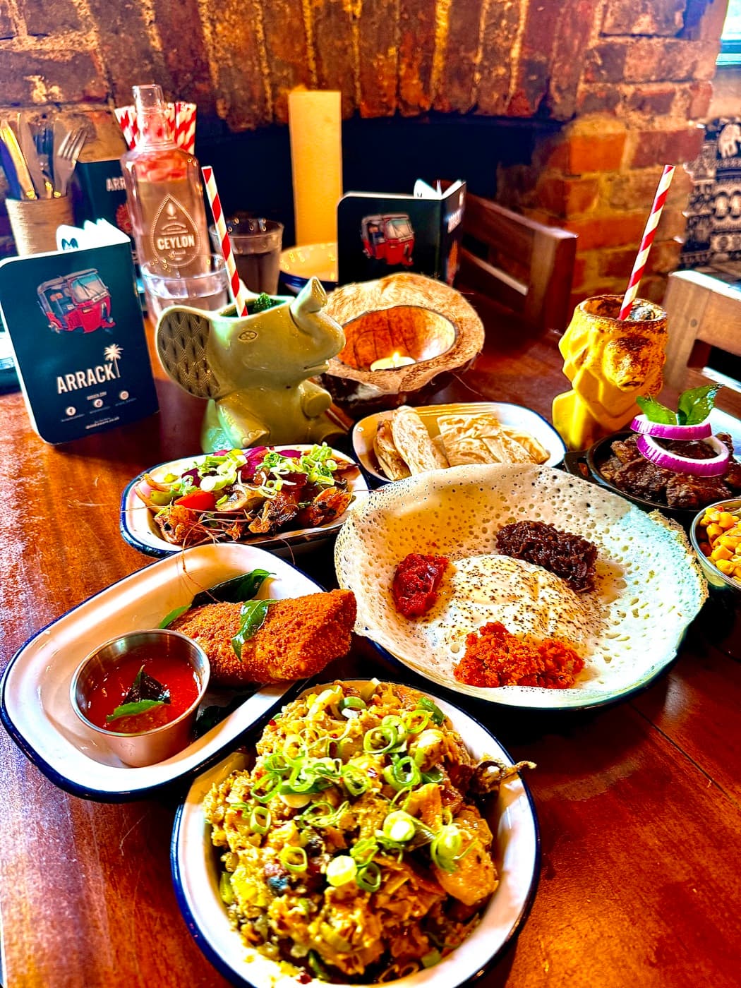 The spread at The Coconut Tree Oxford: an egg hopper with three sambols, mixed seafood kotthu, lamb rolls with dipping sauce, black pork, Sri Lankan sweetcorn and parotta rotti across a wooden table, with cocotails in ceramic elephant and lion mugs behind