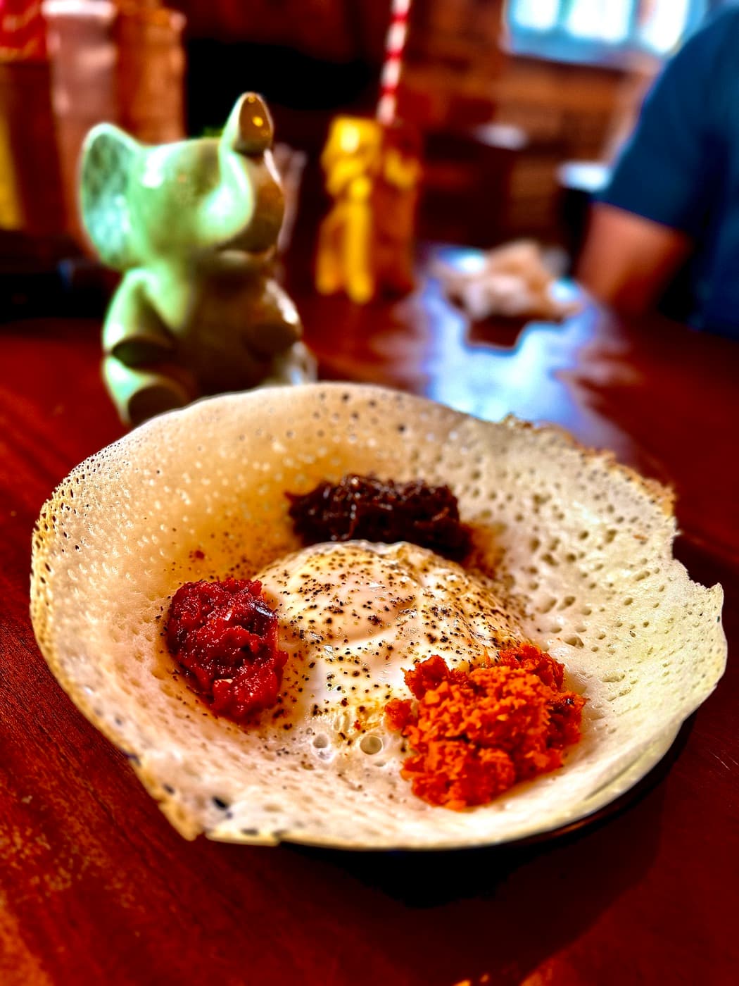 An egg hopper at The Coconut Tree Oxford: a bowl-shaped coconut milk pancake with crisp lacy edges holding a fried egg, ringed by coconut sambol, seeni sambol and lunu miris