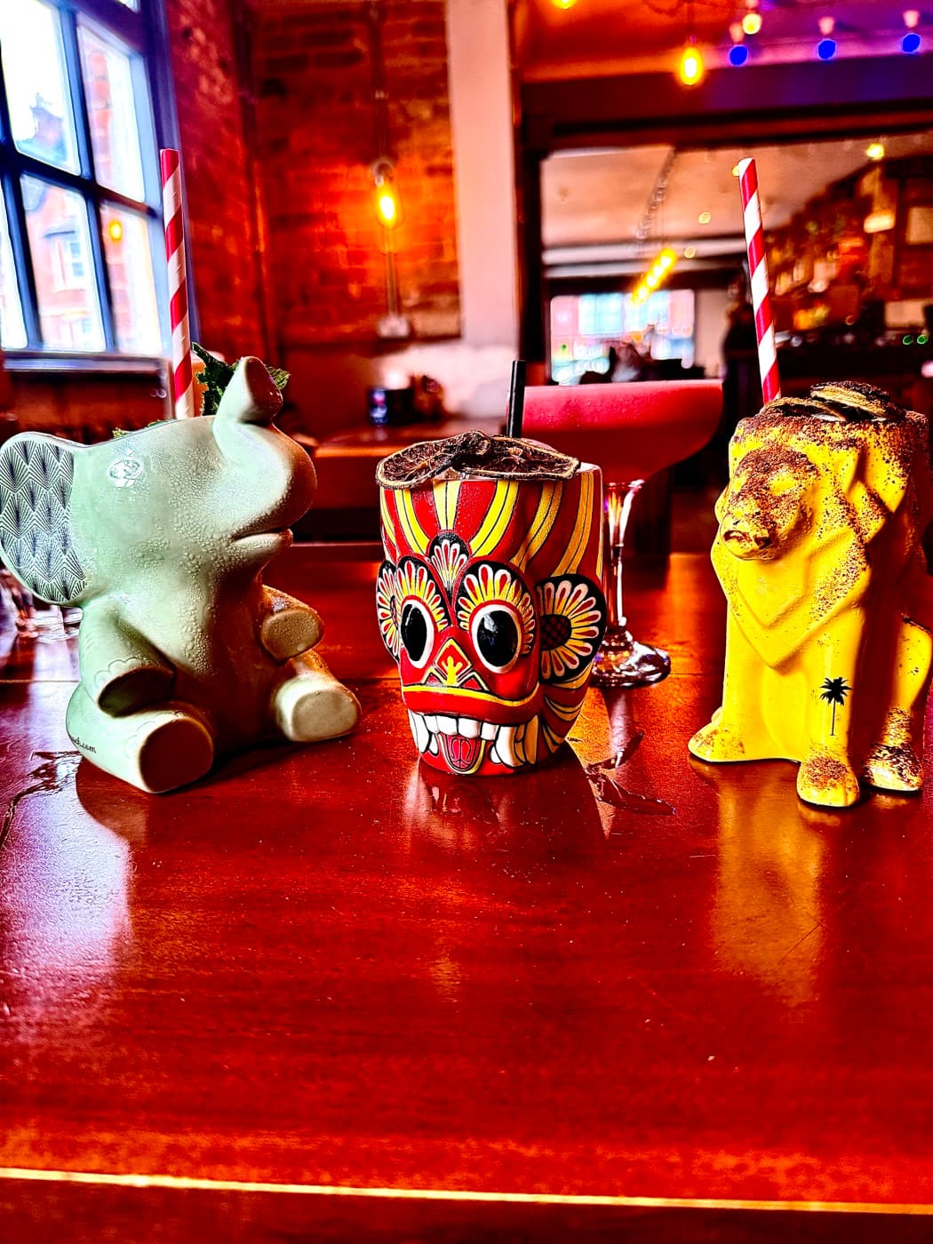 Three cocotails at The Coconut Tree Oxford lined up on the table in ceramic mugs: a pale green elephant, a red and yellow Sri Lankan devil mask and a yellow lion, each with a paper straw