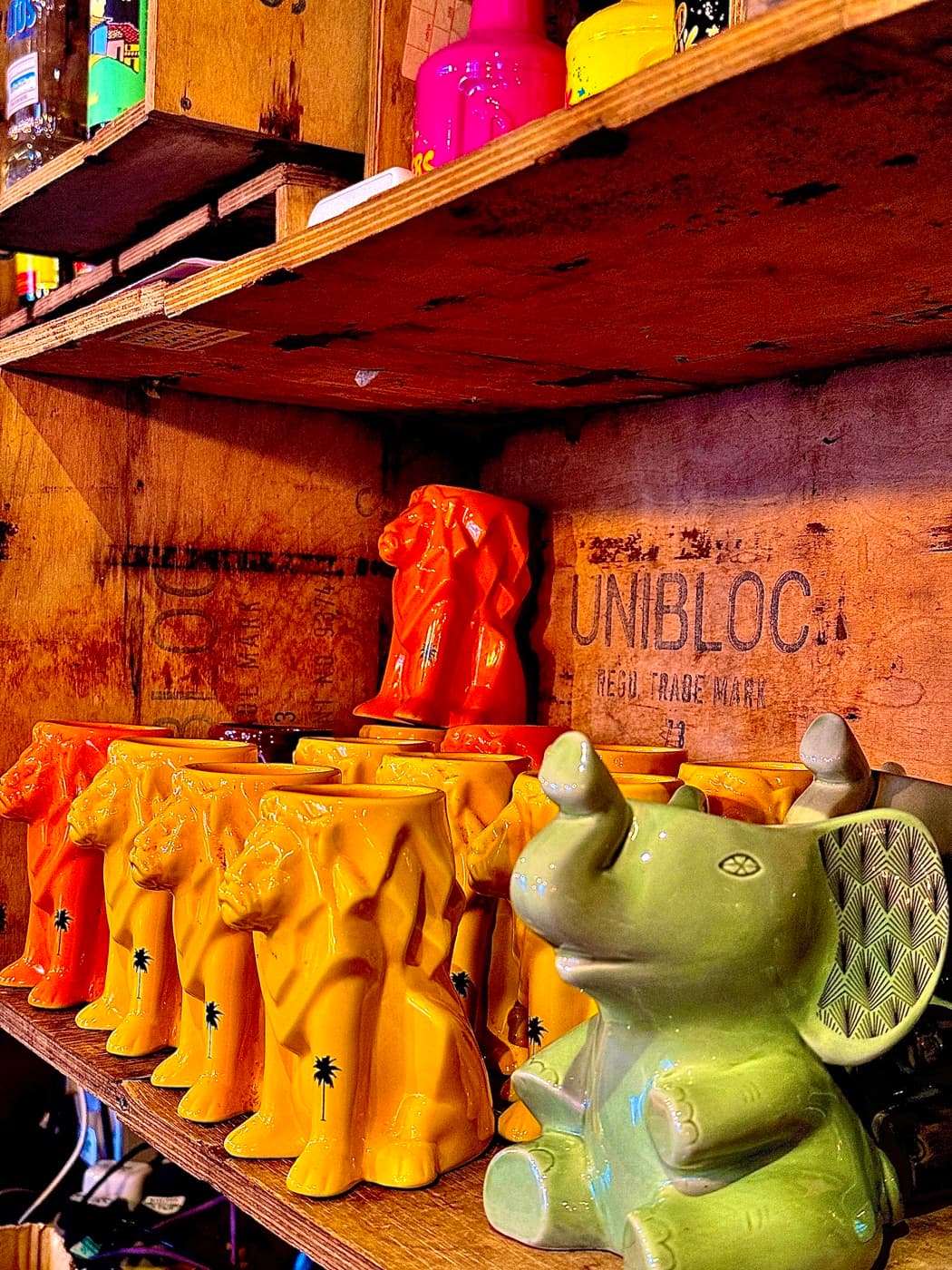 Rows of ceramic cocotail mugs stacked in wooden crates behind the bar at The Coconut Tree Oxford: yellow and orange lion mugs and a pale green elephant mug