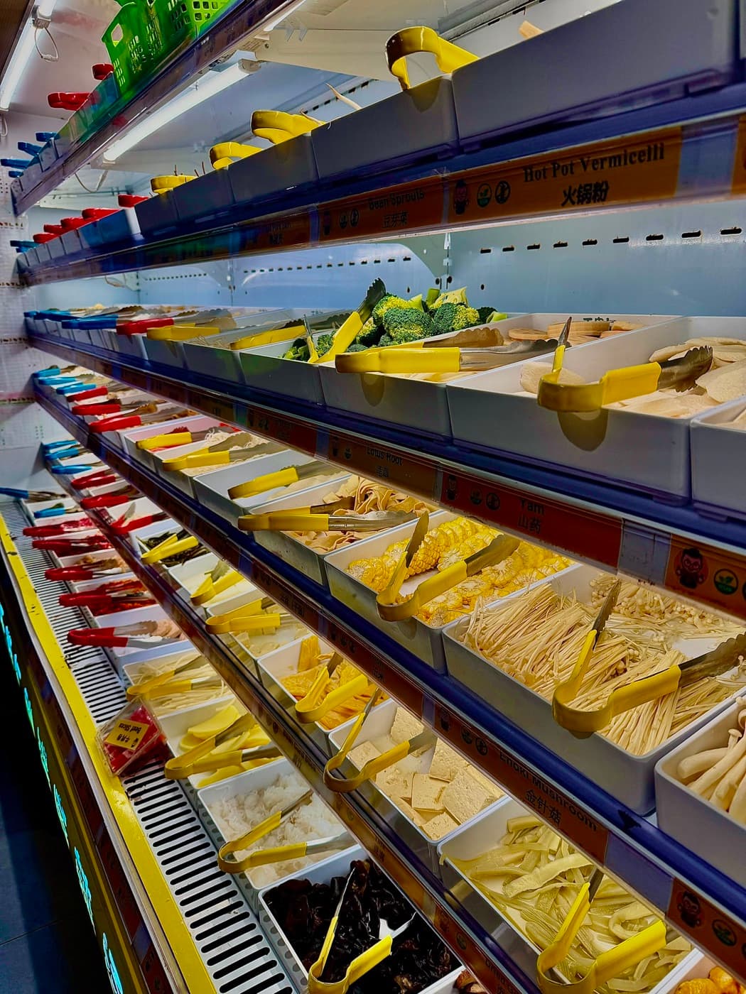 The ingredient display wall at Mr. Wang Hot Pot Oxford, with chilled trays of bean sprouts, hot pot vermicelli, lotus root, yam, sweetcorn, enoki mushrooms, tofu and broccoli, each with its own tongs