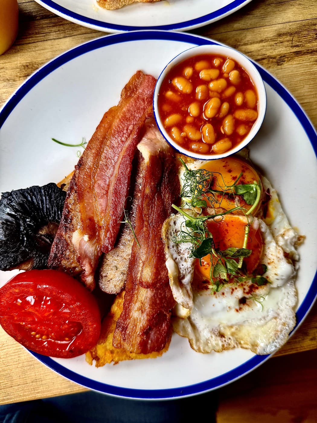 The Full Oxford at Oxford Brunch Bar: streaky bacon, sausage, a flat mushroom, grilled tomato, a hash brown, a bowl of baked beans and two fried Cackleberry Farm eggs on a blue-rimmed plate