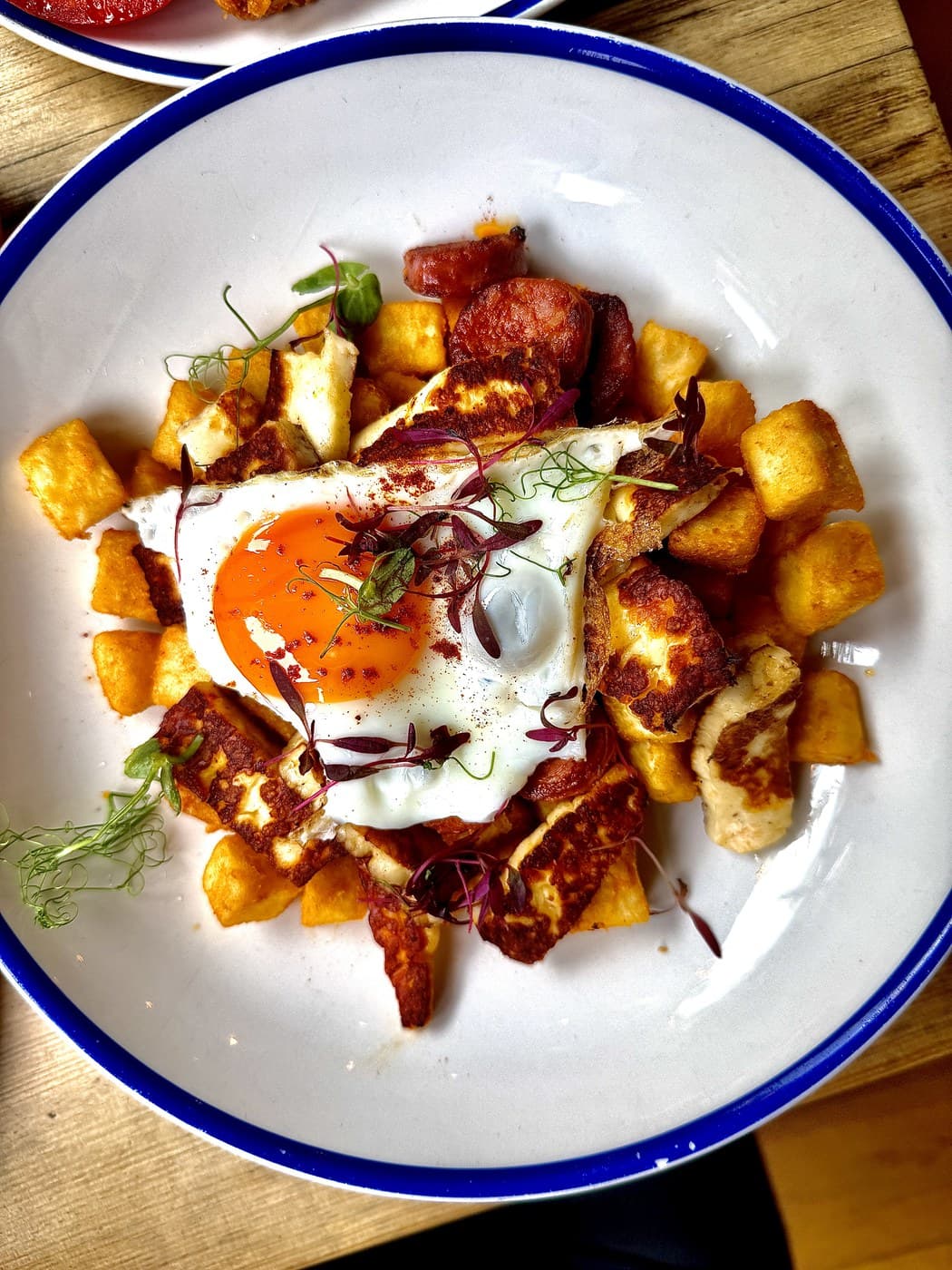 Papa's Plate at Oxford Brunch Bar seen from above: crisp home potatoes topped with grilled chorizo, halloumi and a fried Cackleberry Farm egg with a deep orange yolk, scattered with micro herbs on a blue-rimmed enamel plate