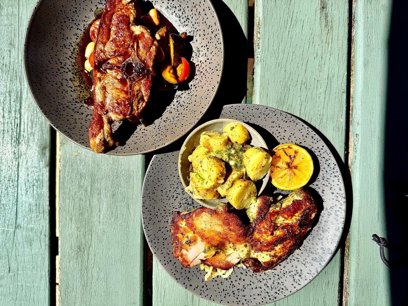 Overhead of two mains in the garden at The Prince of Wales Iffley: a lamb Barnsley chop with butterbeans and tomatoes, and a half grilled chicken with warm potato salad and a charred lemon